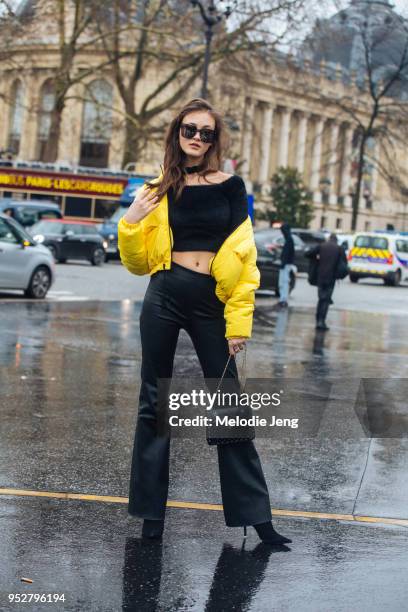 Model Greta Varlese wears a yellow cropped puffer jacket off her shoulders, a black crop top, black wide legged pants, black heels, and a black purse...