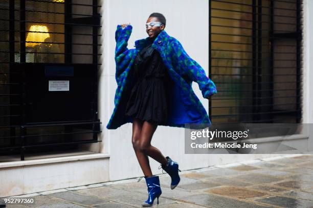 Model Nicole Atieno busts a dance move and wears the dramatic face makeup, a blue and green printed fur coat, blue dress, and blue rain repellent...