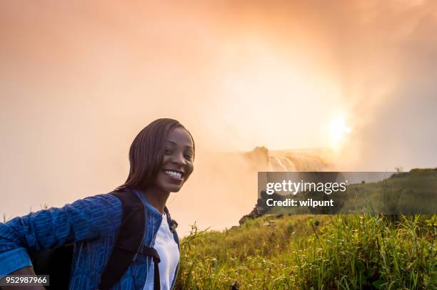 woman with backpack lookingat camera with victoria falls at sunrise - victoria falls stock pictures, royalty-free photos & images