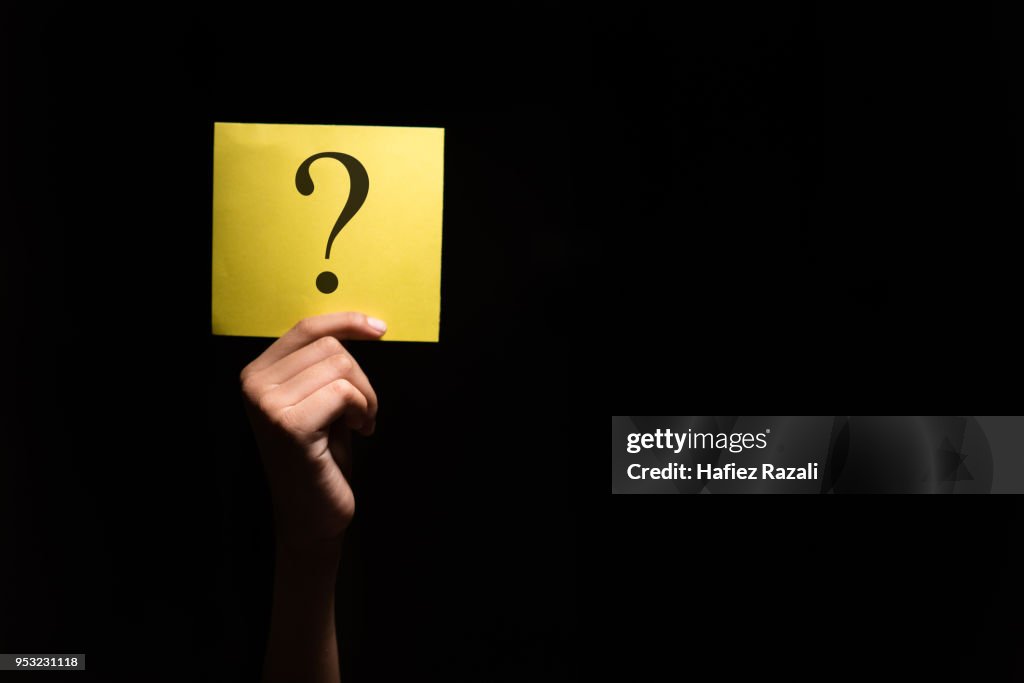 Close-Up da mão segurando ponto de interrogação no papel amarelo contra o fundo preto