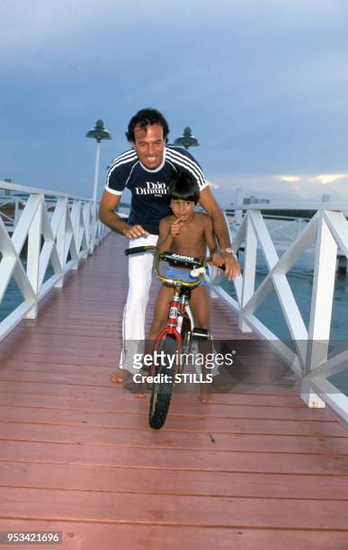 Portrait du chanteur espagnol Julio Iglesias et de son fils sur un vélo sur une jetée près de la mer, circa 1980.