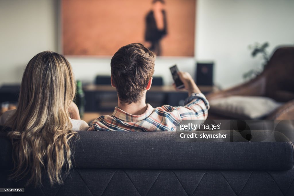 Rear view of a couple relaxing on sofa in the living room and watching a movie on TV.
