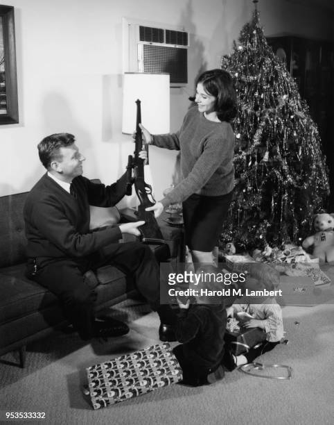 MID ADULT WOMEN GIVING RIFLE TO MAN WITH CHILDREN LOOKING AT THEM AT CHRISTMAS