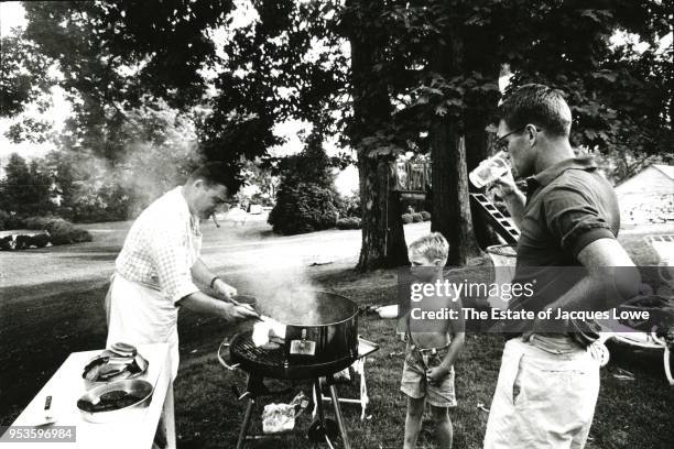 Attorney General Robert F Kennedy drinks from a glass as he, along with a young boy, watch an unidentified man in an apron grills hamburgers on a...