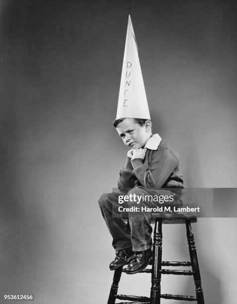 SCHOOLBOY SITTING ON STOOL WITH DUNCE HAT