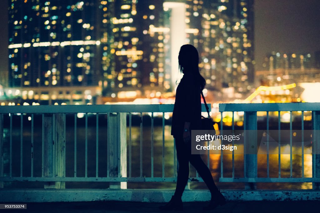 Silhouette of woman walking by the pier overlooking the cityscape at night