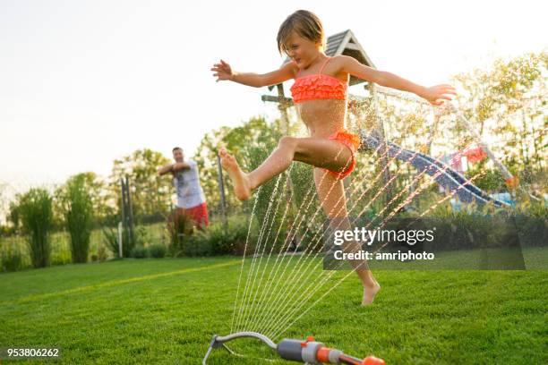 girl jumping over water sprinkler in garden - jumping sprinkler stock pictures, royalty-free photos & images