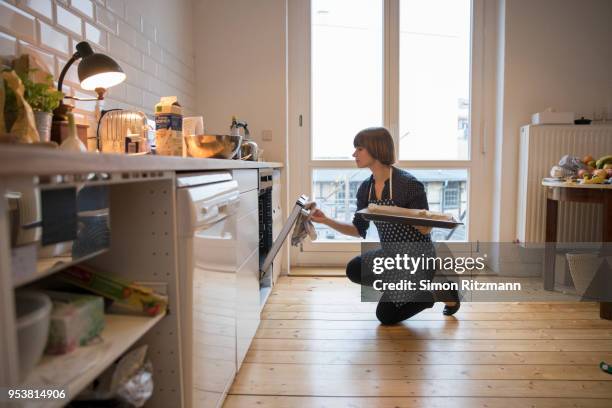 beautiful woman looking in oven while holding baking sheet in kitchen - homemaker stock pictures, royalty-free photos & images