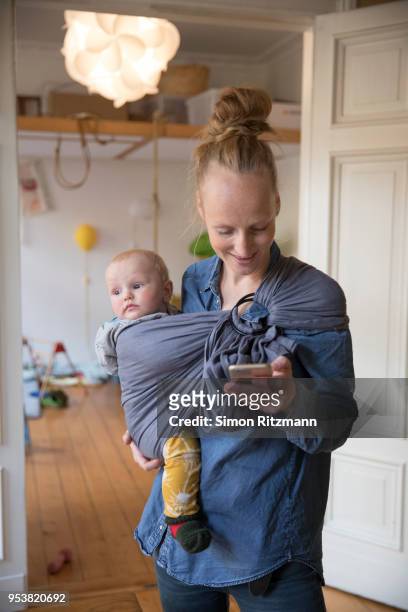 happy mother texting message on cell phone while holding baby in sling - modern boy hipster stock-fotos und bilder