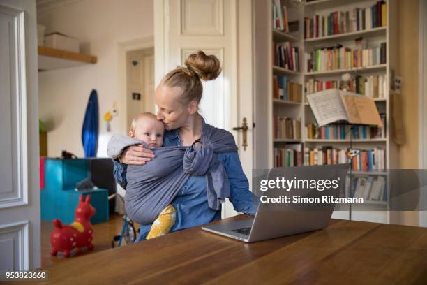 mother holding baby in sling while working at laptop in home office - baby carrier stock pictures, royalty-free photos & images