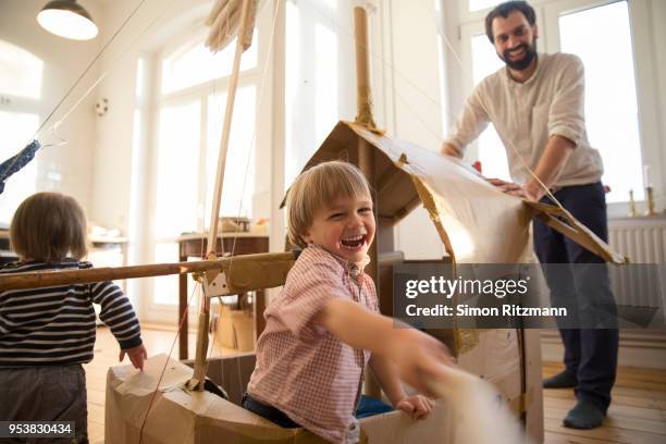 playful son and father playing with cardboard sailingboat at home - sailingboat stock-fotos und bilder