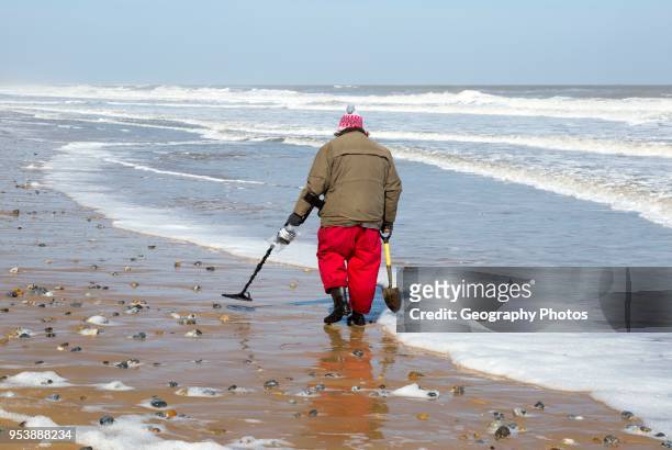 Male detectorist using metal detector walking along sandy beach Hemsby, Norfolk, England, UK.