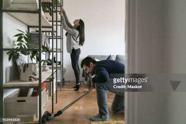 man vacuuming floor while woman cleaning shelf at home - arrangiare foto e immagini stock