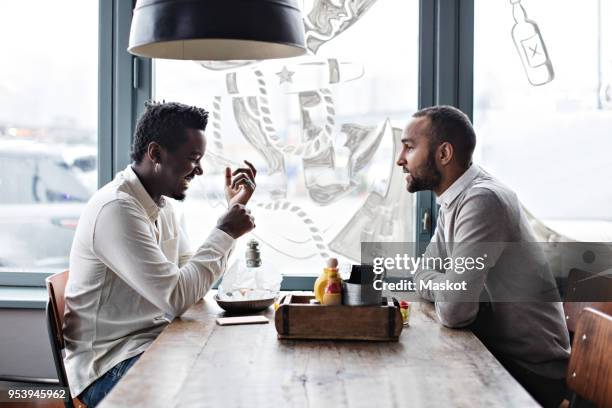 side view of smiling friends talking while having brunch by window at restaurant - amigos hombres en restaurant fotografías e imágenes de stock