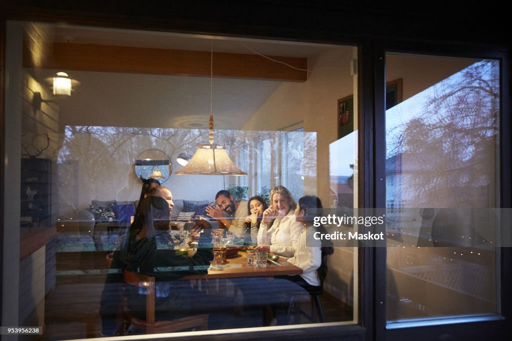 Happy family having dinner at table seen through glass window during sunset