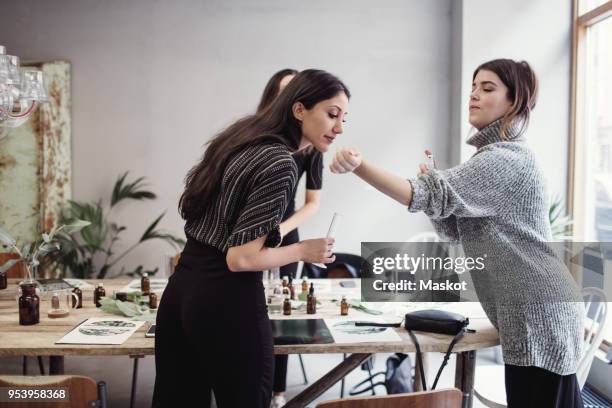 young woman smelling perfume fragrance from female colleagues wrist while standing at workshop - senses stock pictures, royalty-free photos & images