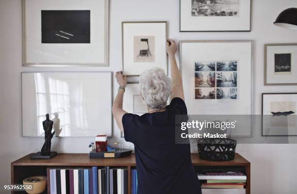 rear view of retired senior woman adjusting picture frame on wall over bookshelf at home - adjusting ストックフォトと画像