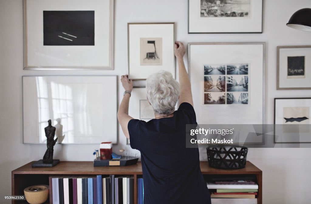 Rear view of retired senior woman adjusting picture frame on wall over bookshelf at home