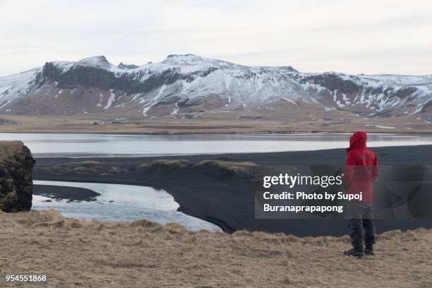 tourist in red jacket looking at view of snowcapped mountain part of myrdalsjokull glacier at kirkjufjara beach,south iceland - myrdalur stock pictures, royalty-free photos & images