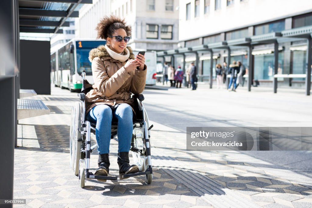 Gehandicapte vrouw in een rolstoel te wachten op een busstation en het gebruik van slimme telefoon
