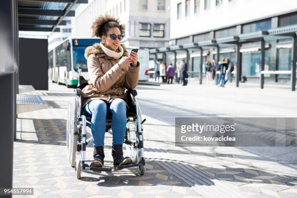 disabled woman in a wheelchair waiting at a bus station and using smart phone - wheelchair access stock pictures, royalty-free photos & images