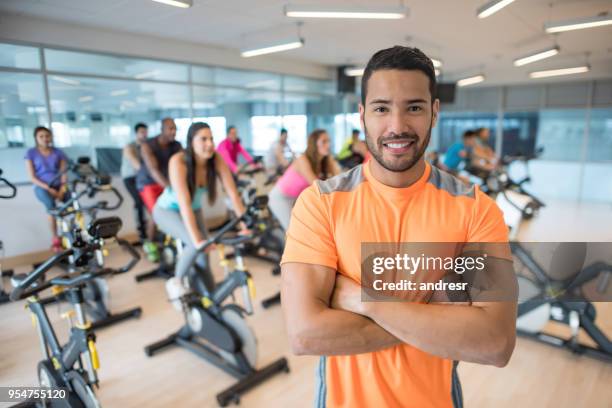 portrait of handsome exercising instructor looking at camera with arms crossed and class behind him on the stationary bikes - exercise instructor stock pictures, royalty-free photos & images
