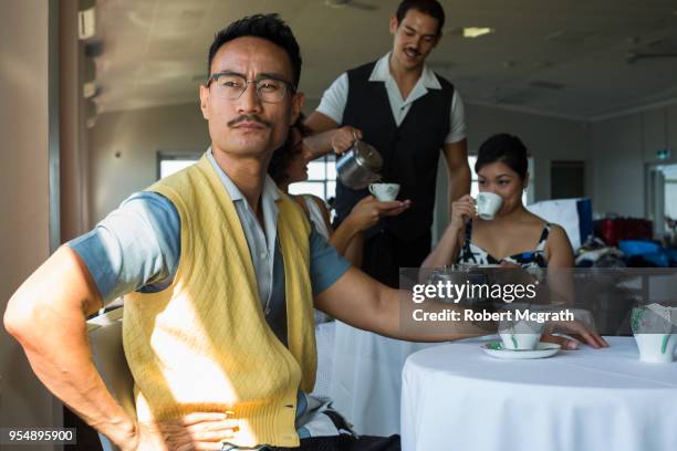 asian man with moustache looks contemptuously out the window while his female companions enjoy their morning tea served by a european waiter. - robert mcgrath stock-fotos und bilder