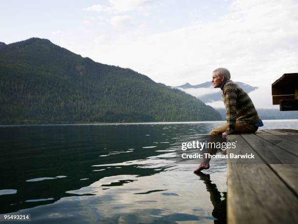 man sitting on edge of dock with feet in water - bergsteiger stockfoto's en -beelden