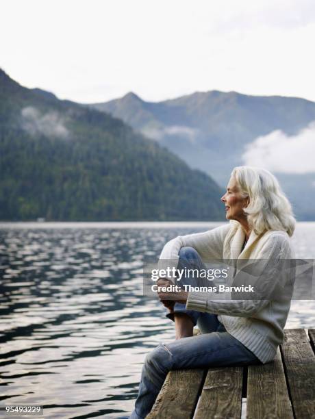 mature woman sitting on edge of dock on lake - waters edge imagens e fotografias de stock