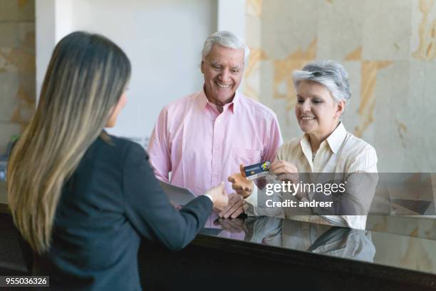 beautiful senior couple doing check in at hotel and senior woman handing a loyalty rewards card to receptionist all looking very happy - travel loyalty stock pictures, royalty-free photos & images