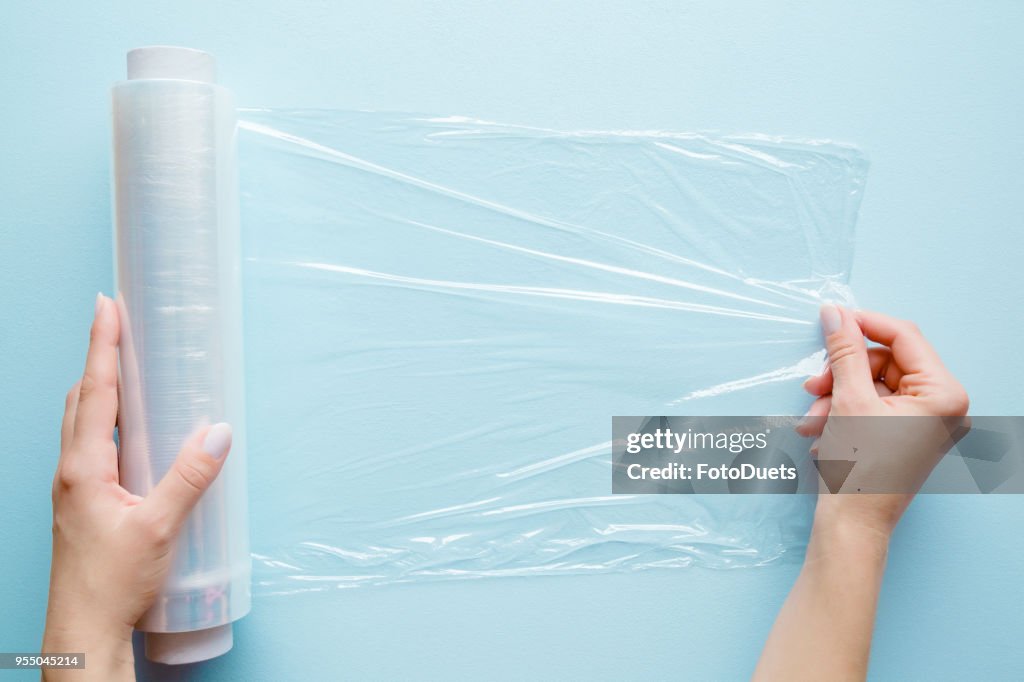 Woman's hand using a roll of transparent polyethylene food film for packing products on the pastel blue table. Empty place for text or logo.