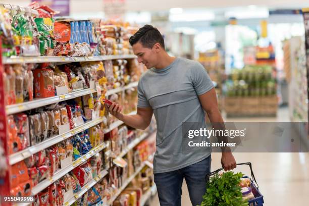 joven solo comprando víveres en el supermercado leyendo la etiqueta de un producto de aspecto muy feliz - pasillo objeto fabricado fotografías e imágenes de stock