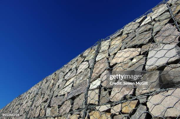 stone retaining wall encased in wire mesh aginst a clear blue sky - stützmauer stock-fotos und bilder