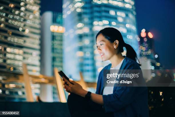 beautiful young woman using cell phone on urban balcony overlooking illuminated city skyline of hong kong at night - corporate skyline stock pictures, royalty-free photos & images