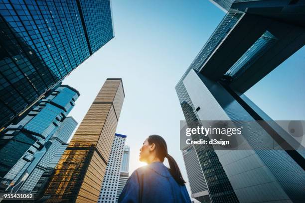 low angle view of woman standing against modern skyscrapers and blue sky in city at sunrise - hong kong business photos et images de collection