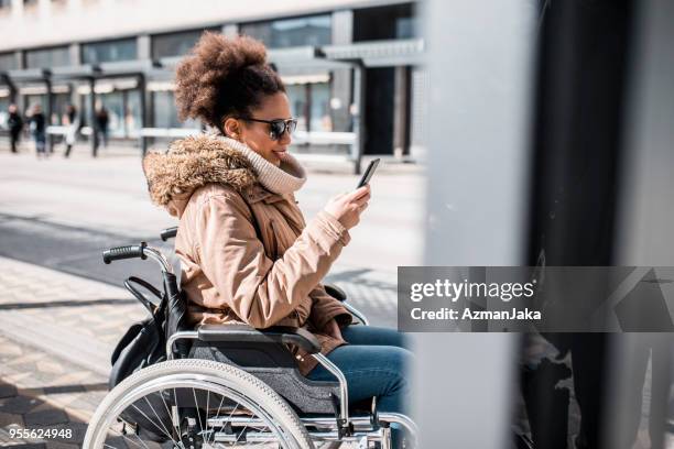 behinderte frau im rollstuhl mit smartphone und warten auf einen bus - barrierefrei stock-fotos und bilder