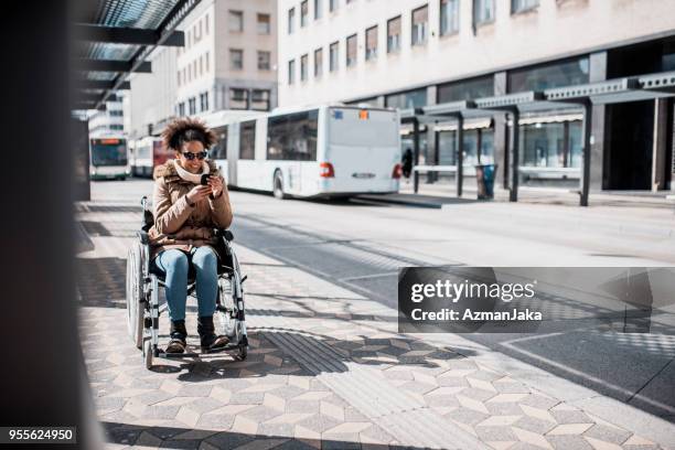 disabled woman in wheelchair using smart phone and waiting for a bus - wheelchair ramp stock pictures, royalty-free photos & images