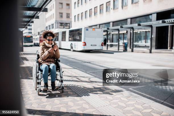 disabled woman in wheelchair using smart phone and waiting for a bus - waiting bus stock pictures, royalty-free photos & images