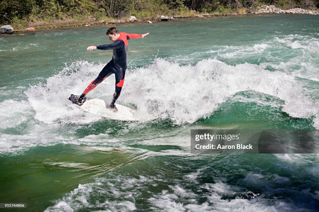 Man in a red wetsuit standing on surfboard surfing a greenish wave in the middle of a river.