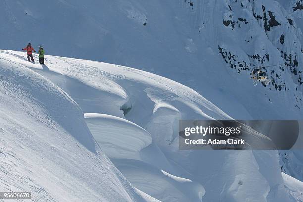 two skiers waiting to ski with a helicopter in the background in alaska. - heli skiing stock pictures, royalty-free photos & images