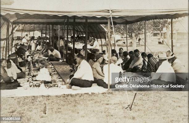 Photograph of an Osage election dinner, early twentieth century. The dinner is outdoors under tents, and people are gathered in rows with food laid...