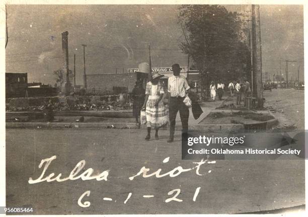 An African American couple walking across a street with smoke rising in the distance after the Tulsa Race Massacre, Tulsa, Oklahoma, June 1921.