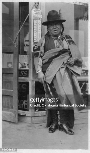 Photograph of an unidentified Osage man standing on the sidewalk outside of a retail shop, Fairfax, Oklahoma, early twentieth century. He is wearing...
