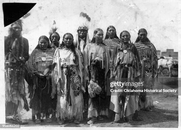 Photograph of a group of nine Kiowa dancers wearing fringed buckskins with beaded embellishments, shells and beaded leather moccasins, early...