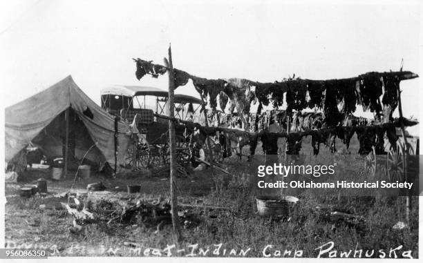 Photograph of Osage Indians drying bison meat at their camp, near Pawhuska, Oklahoma, early twentieth century. A horse-drawn carriage and tent are in...