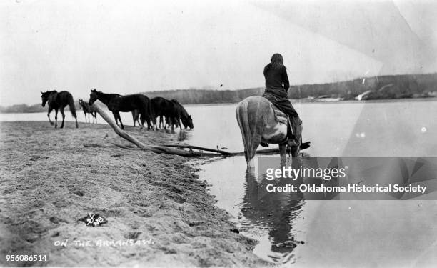 Photograph of an Osage man on the Arkansas River, between 1910 and 1918. The man is sitting on a horse in the water with horses in the background on...