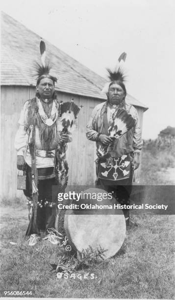 Photograph of Osage George Newalla and an unidentified Osage man with a drum at their feet, early twentieth century. The men are wearing roach...