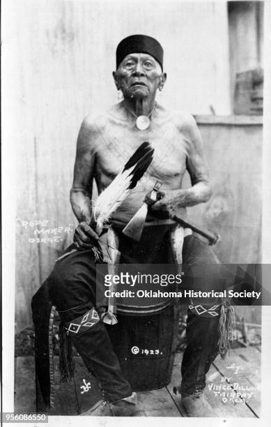 Photograph of an Osage Rope Maker seated on a barrel holding a hatchet and a feathered fan, 1923. He is wearing pants with beaded embellishments, no...