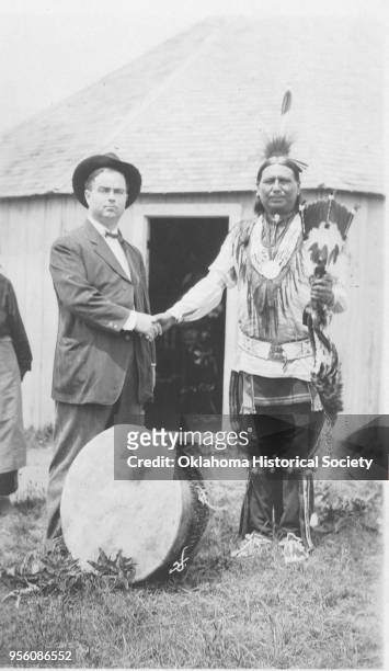 Photograph of Osage George Newalla who is shaking hands with an unidentified in a suit and hat in front of a roundhouse, 1910. There is a large drum...