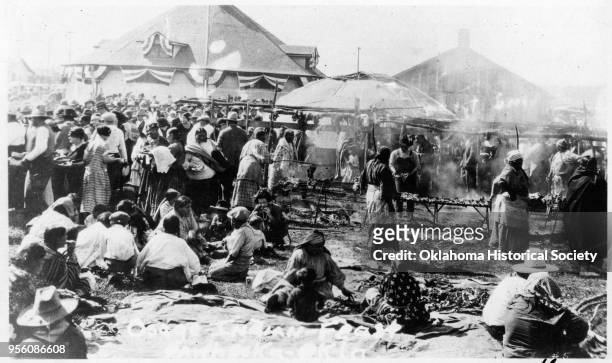 Photograph of a large crowd of people at an Osage feast, Pawhuska, Oklahoma, early twentieth century. There are people cooking meat on a grate over...